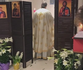 An Orthodox priest stands at the Holy Table inside the altar during Divine Liturgy, surrounded by icons and liturgical furnishings.