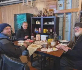 Parishioners and clergy sit together at a table sharing food and conversation during fellowship after an Orthodox service.